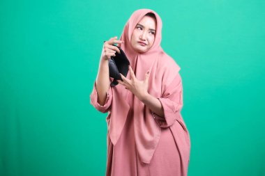 Boş bir cüzdan tutarken sıkıntılı görünen tesettürlü bir kadın, yeşil arka planda izole edilmiş mali zorlukları ve ekonomik zorlukları sembolize ediyor..