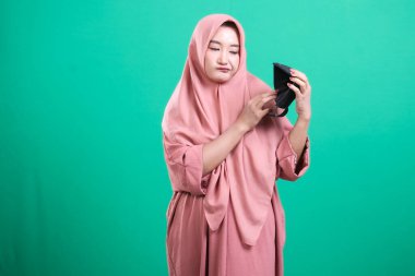 Pembe tesettürlü Asyalı bir kadının portresi, yeşil arka planda izole edilmiş, siyah koruyucu yüz maskesi takıyor. Sağlık sorunları yüzünden hayal kırıklığına uğramış görünüyor..