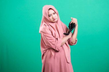 Boş cüzdanını gösteren tesettürlü, hayal kırıklığına uğramış Müslüman bir kadın. Ekonomik zorlukları ya da yoksulluğu sembolize ediyor. Yeşil arka planda izole edilmiş..