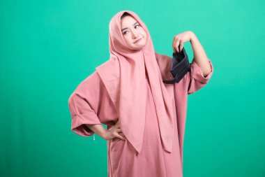 Mutsuz, tesettürlü, boş bir cüzdan gösteren Müslüman kadın. Portre, yeşil arka planda izole edilmiş mali zorlukları tasvir ediyor. Modern bir birey için ekonomik zorlukları ve kişisel para sorunlarını gözler önüne seriyor.