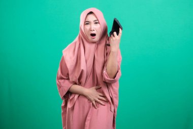 Başörtüsü takan şok olmuş Müslüman bir kadın, yeşil arka planda izole edilmiş mali zorlukları sembolize ederek elinde boş bir cüzdan tutarken inanmadığını ifade ediyor. Gelir ve harcama zorlukları konusunda sıkıntı ve belirsizlik gösteriyor..