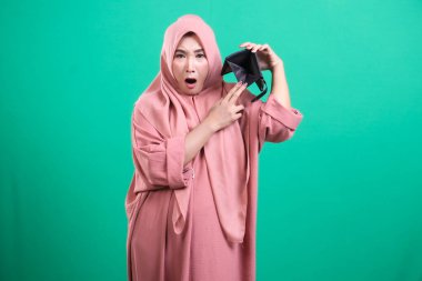 Boş bir cüzdan taşıyan ve yeşil arka planda izole edilmiş, başörtüsü takan şok olmuş bir kadın. Finansal sorunları, ekonomik durgunluğu ve yeşil arka planda izole edilmiş fon eksikliğini simgeliyor.