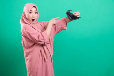 Boş bir cüzdan tutan tesettürlü Müslüman bir kadın, yeşil arka planda izole edilmiş mali istikrarsızlık endişesini yansıtıyor. Onun ifadesi ekonomik krizlerin etkilerini net bir şekilde yakalıyor..