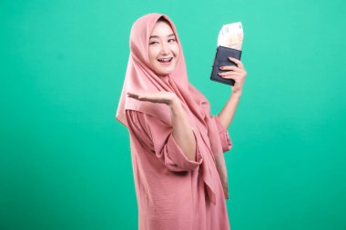 Mutlu genç Müslüman kadın, neşeli bir ifadeyle para dolu cüzdanını gösteriyor, zenginliği resmediyor, yeşil bir arka planda izole edilmiş. Başarı göstergesi olarak, elinde para tutuyor ve poz veriyor..