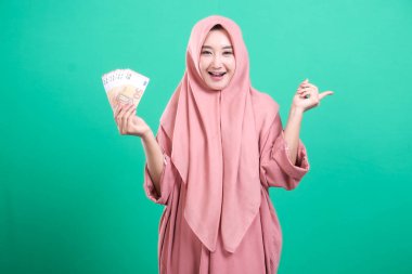 Tesettürlü neşeli bir kadın, elinde avro banknotları ve boş bir yeri işaret ediyor. Yeşil arkaplanda izole edilmiş bir sunum gösteriliyor.