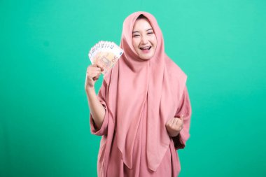 Başörtüsü takan neşeli Müslüman bir kadın, mali başarısını yeşil bir arkaplanda izole bir şekilde elinde Euro banknotları tutarak kutluyor. Mutlu ifadesi memnuniyetini gösteriyor..