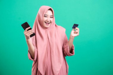 Mutlu Müslüman bir kadın, yeşil bir arka planda izole edilmiş, kredi kartını ve cep telefonunu göstererek başarılı bir çevrimiçi işlemi kutluyor..