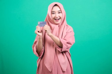 Tesettürlü genç Müslüman kadın elinde banka kartı tutarak para transferleri hakkındaki iyimserliğini dile getiriyor. Yeşil bir arkaplanda, modern finans dünyasında güven ve başarı yayıyor..