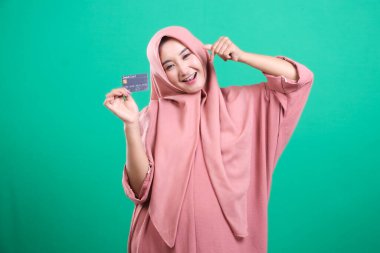 Tesettürlü neşeli genç Müslüman kadın, son moda gül kıyafetleri içinde zahmetsiz ödemeler için bir kredi kartı tutuyor. Yeşil arkaplanda izole edilmiş.