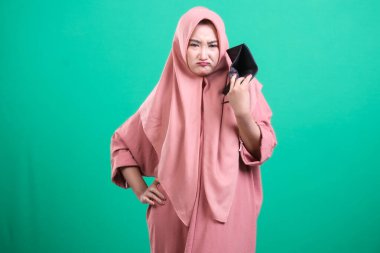 Ümitsiz görünen ve boş cüzdanını tutan genç bir kadın, yeşil arka planda izole edilmiş mali sorunlarla karşı karşıya. O, tesettürlü Müslüman bir kadın. Ekonomik umutsuzluğunu dile getiriyor..