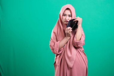 Başörtüsü takan üzgün genç bir kadın üzgün bir ifadeyle boş cüzdanını gösteriyor. Yeşil arka planda izole edilmiş mali sıkıntıları temsil ediyor. Para sorunlarını tasvir ediyor..