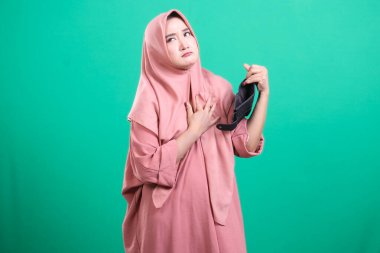 Başörtüsü takmış üzgün bir Müslüman kadın, yeşil bir arka planda izole edilmiş, yoksulluğu ve parasızlığı gösteren boş bir cüzdan tutuyor. Bu görüntü finansal sorunları ve umutsuzluğu iletir.