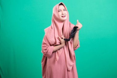 Türbanlı bir Asyalı kadın, yeşil bir arka planda izole edilmiş nefes darlığı nedeniyle maskesini çıkarırken rahatsızlık ve rahatlama belirtileri gösteriyor. Bu, uzun süreli maske takmanın getirdiği yorgunluğu vurgular..