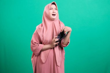 Başörtüsü içindeki sıkıntılı kadın boş bir cüzdan gösteriyor, mali krizi ve ekonomik sıkıntıyı simgeliyor, yeşil bir arka planda izole edilmiş umutsuzluğu taşıyor. Bir kadın, ekonomik kriz ve kaybın yaşandığı dönemde yoksullukla mücadele ediyor.