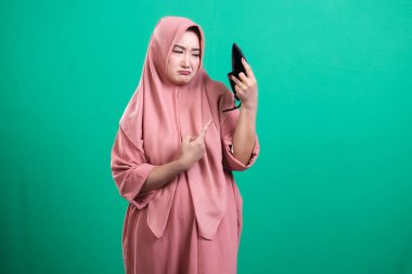 Hüsrana uğramış Müslüman kadın boş cüzdanını işaret ederek parasal zorlukları ve parasal sıkıntıyı sembolize ediyor, yeşil bir arka planda izole edilmiş. Ekonomik durumu kötü.