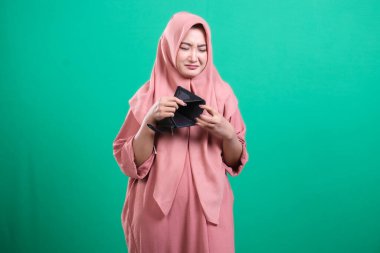 Endişeli, tesettürlü Müslüman bir kadın, boş cüzdanına bakıyor, mali sıkıntıyı simgeliyor, yeşil arka planda izole edilmiş sınırlı kaynakların stresini tasvir ediyor..