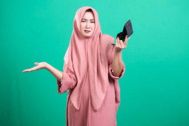Başörtüsü olan üzgün bir Müslüman kadın, yeşil arka planda izole edilmiş mali zorlukları vurgulayarak boş cüzdanını gösteriyor. Finansal durumu onu hayal kırıklığına uğrattı..