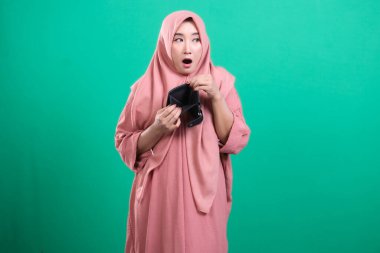 Yeşil bir arka planda izole edilmiş, harcamalar nedeniyle mali sıkıntı gösteren boş bir cüzdan tutan Müslüman kadın şok oldu. Para yönetimi ve finansal zorlukları simgeliyor..