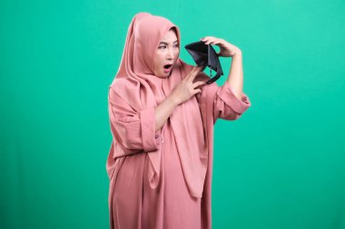 Başörtüsü takmış endişeli bir Müslüman kadın yeşil bir arka planda izole edilmiş hayal kırıklığına uğramış bir ifadeyle boş cüzdanına bakıyor. Bu vitrinler finansal zorlukları gösteriyor..