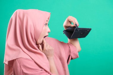Yeşil arka planda izole edilmiş, maddi sıkıntıların olumsuz etkilerini gösteren, boş bir cüzdana bakan tesettürlü bir kadının portresi..