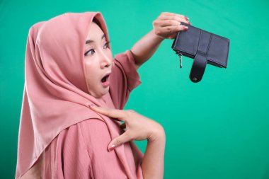 Genç Müslüman bir kadın endişeli bir bakışla boş cüzdanını gösteriyor, finansal zorlukları yansıtıyor, yeşil bir arka planda izole edilmiş, mali endişeyi ve yoksulluğu gösteriyor..