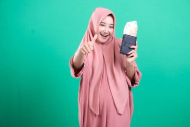 Heyecanlı tesettürlü Müslüman kadın para dolu bir cüzdan sergiliyor ve yeşil bir arka planda kendini neşeli, izole edilmiş hissediyor. Mali başarı ve bolluk gösteriliyor.