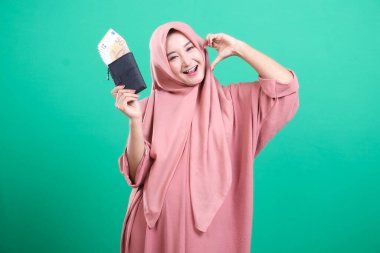 Para dolu bir cüzdan gösteren tesettürlü neşeli Müslüman bir kadının portresi ve yeşil bir arka planda izole edilmiş, parmaklarıyla kalp işareti yapan. Mali başarı ve olumlu yatırım sonuçları gösteriliyor.