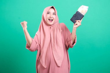 Başörtüsü takan heyecanlı bir Müslüman kadın, para ve yeşil arka planda izole edilmiş neşeli bir ifadeye sahip bir cüzdan tutarak mali başarısını kutluyor..