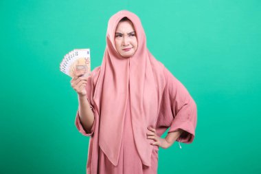 Pembe tesettürlü düşünceli bir Müslüman kadın, yeşil bir arka planda izole edilmiş, şüpheci bir bakışla Avrupa banknotları tutuyor. Bu resim finansal temaları temsil eder.