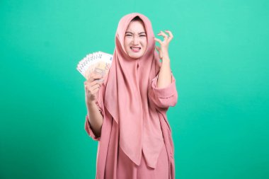 Avro banknotları tutan tedirgin genç Müslüman kadın, mali ve para yönetimi konusunda stres gösteriyor, yeşil bir arka planda izole edilmiş.