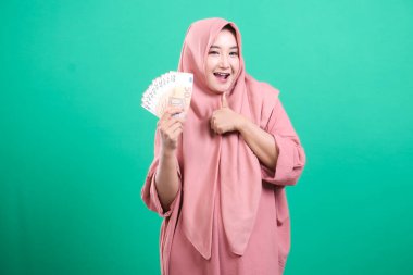 Başörtüsü takan memnun bir Müslüman kadın, Avrupa banknotlarını elinde tutuyor ve yeşil bir zemine izole edilmiş neşe ve başarı saçan bir onay veriyor. Bu görüntü finansal refahı ve refahı yakalar..