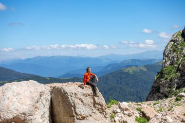 Turuncu kazaklı bir adam yüksek dağların arka planında bir taşın üzerinde oturur.