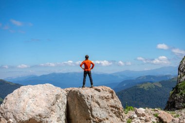 Turuncu kazaklı bir adam yüksek dağların arka planında bir taşın üzerinde oturur.