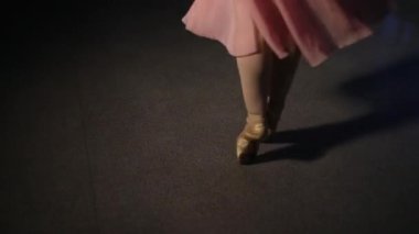 Karanlık bir sahnede ya da stüdyoda egzersiz yapan bir balerinin yakın çekimi. Sivri uçlu ayakkabılı kadın bacakları. Balerin klasik bale adımlarını gösteriyor. Yavaş çekim