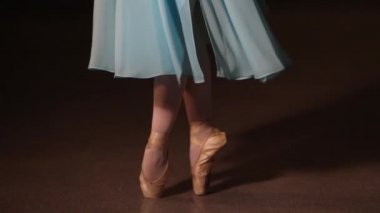 Karanlık bir sahnede ya da stüdyoda egzersiz yapan bir balerinin yakın çekimi. Sivri uçlu ayakkabılı kadın bacakları. Balerin klasik bale adımlarını gösteriyor. Yavaş çekim