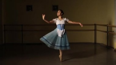 Koyu renk bir bale sınıfında dans eden moda balerini. Kız sahne kostümüyle dans figürleri sergiliyor. Yavaş çekim.