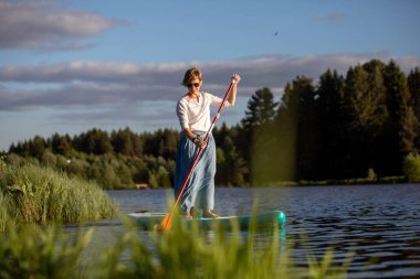 Etekli genç bir kız orman göletinde SUP sörf tahtasına biniyor.