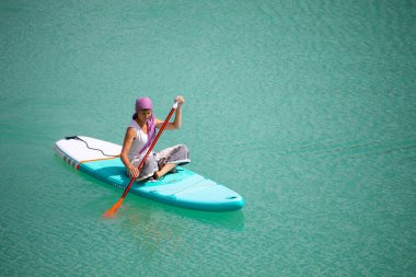 Elbiseli bir kız parlak turkuaz suyla dolu bir gölette sallanan bir tahtanın üzerinde süzülüyor. Seyahat için sıcak bir yaz günü. Bir kuadkopterin üst görüntüsü. Hava fotoğrafçılığı