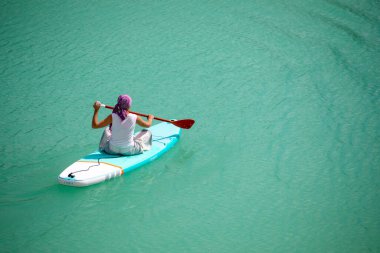Elbiseli bir kız parlak turkuaz suyla dolu bir gölette sallanan bir tahtanın üzerinde süzülüyor. Seyahat için sıcak bir yaz günü. Bir kuadkopterin üst görüntüsü. Hava fotoğrafçılığı