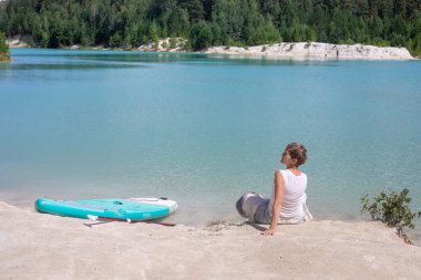 Göl kenarındaki beyaz kumların üzerinde bir kız oturuyor. Gök mavisi turkuaz suyla. Yanında bir glan tahtası var. Yaz tatilleri ve seyahatler