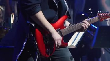 Müzisyen sahnede vücut açılı parlak kırmızı elektro gitar çalar. Sahne ışıklandırması, enerjik parıltı artırıcı atmosfer yaratıyor.