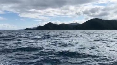 Bereketli yeşil dağlar, bulutlu bir gökyüzünün altında derin mavi okyanus dalgaları ve hareket eden bir tekneden gelen deniz meltemi ile panoramik bir kıyı şeridi.