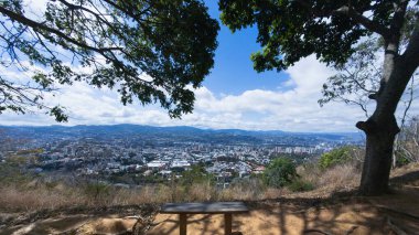 Ağaçların altındaki ahşap bir bank Caracas şehrinin panoramik manzarasını sunuyor, Venezuela, doğa ve mavi gökyüzü tarafından çerçevelenmiş modern bir şehir.
