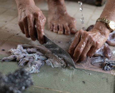Yerli bir insanın fayanslı zeminde taze mürekkep balıklarını temizlemek ve dilimlemek için bıçak kullanırken çekilmiş otantik bir fotoğrafı. Geleneksel deniz ürünleri hazırlama süreci.