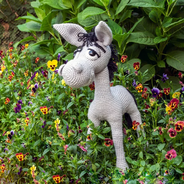 Handmade soft toy amigurumi gray donkey among the greenery of the garden in summer.