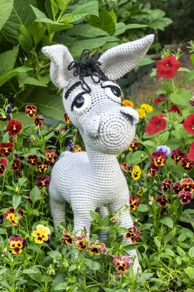 Handmade soft toy amigurumi gray donkey among the greenery of the garden in summer.