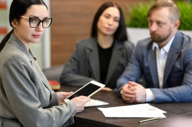 Profesyonel emlakçı elinde dijital tablet tutarken çiftlere modern ofiste emlak anlaşmasıyla ilgili danışmanlık yapıyor. Emlak alım satımı, sözleşme gözden geçirme ve iş görüşmesi kavramı.