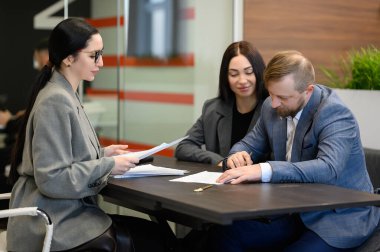 Emlakçı, emlak alım sürecinde çifte yardım ediyor. Kadın modern ofis masasında belgeleri incelerken erkek ev satın alma sözleşmesi imzalıyor. Gayrimenkul işlemleri, yasal evrak işleri ve konut anlaşması kavramı.