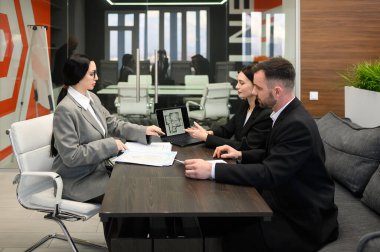 Profesyonel emlakçı modern ofiste emlak danışmanlığı sırasında genç çifte daire planı sunuyor. Müşteriler ev satın alma, ipotek ve konut yatırımlarını tartışırken dizüstü bilgisayardaki düzeni gözden geçiriyorlar