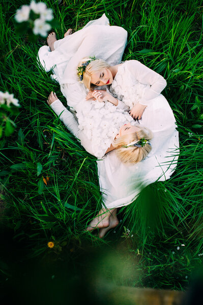 Two blond twins sisters in white dresses lying on green grass